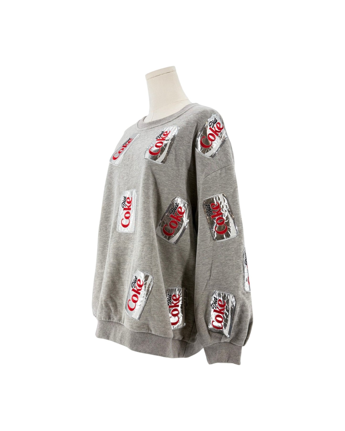 A heather gray crewneck sweatshirt displayed on a mannequin torso, featuring an all-over print of red and white Coca-Cola logos on silver can-like patches.