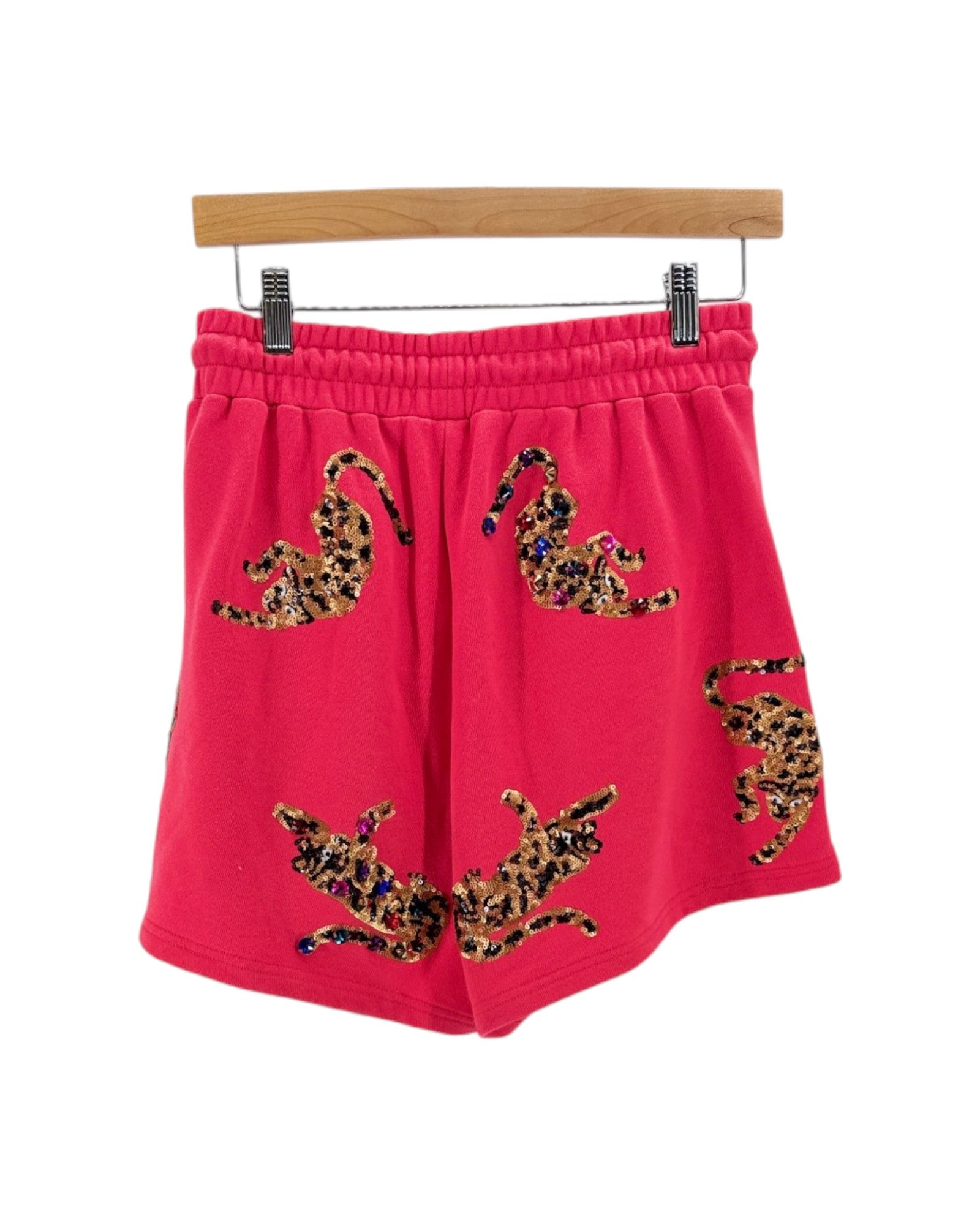 Bright pink sweat shorts with leopard print patches, hanging from a wooden hanger against a white background.