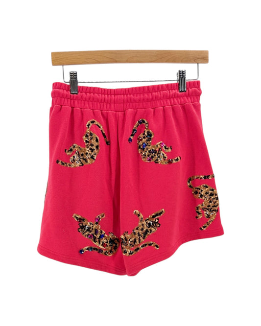 Bright pink sweat shorts with leopard print patches, hanging from a wooden hanger against a white background.