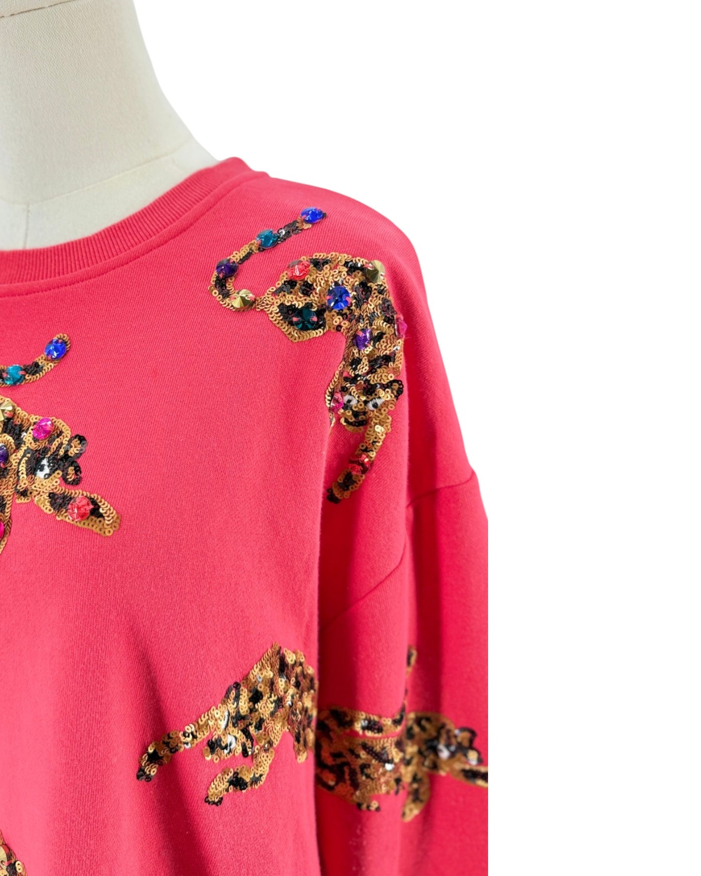 A close-up of a bright pink crewneck sweatshirt on a mannequin, featuring leopard print sequin patches embellished with colorful rhinestones on the shoulders and sleeves.