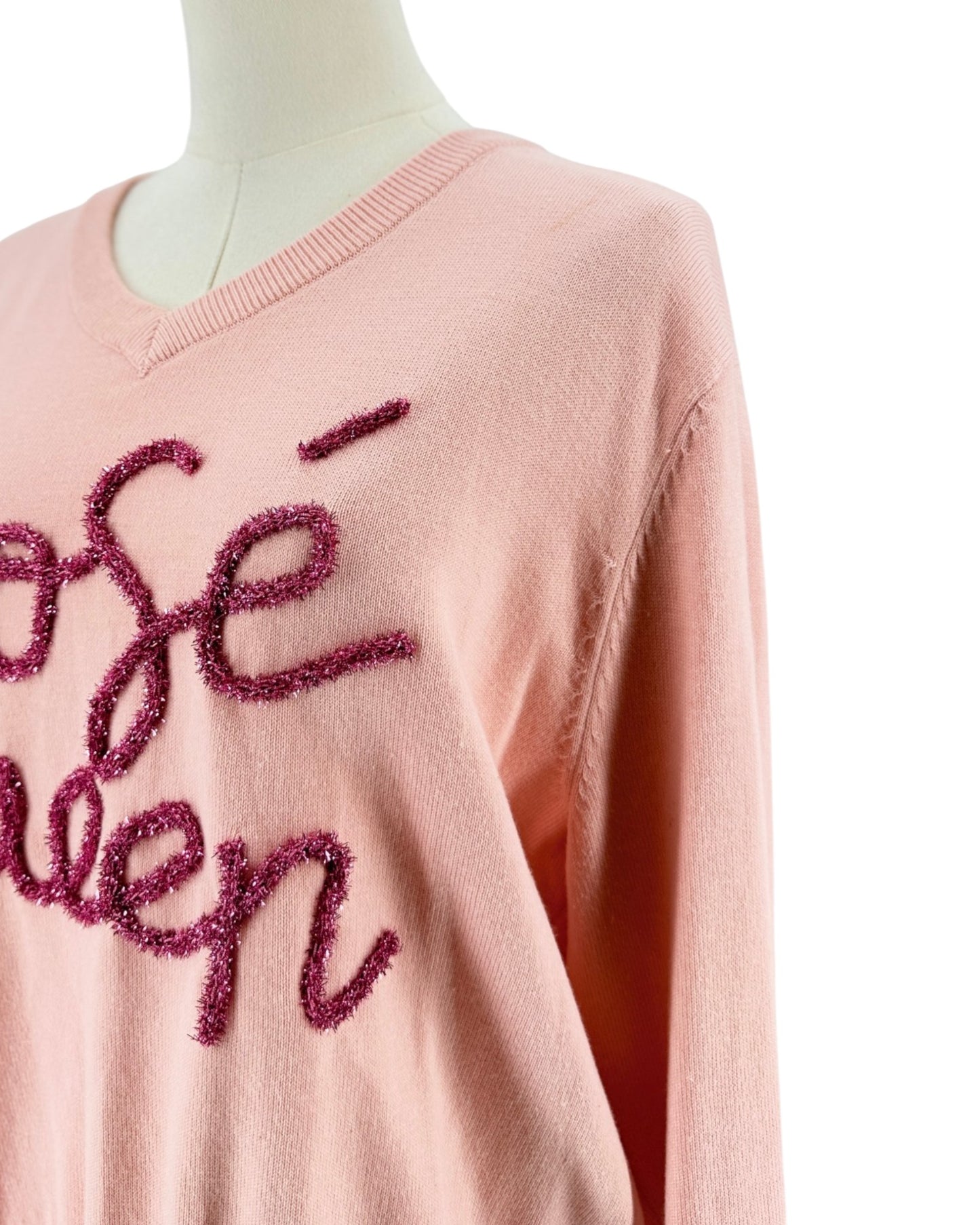 A close-up of a light pink knit sweater displayed on a mannequin, featuring textured, glittery magenta cursive lettering on the front.