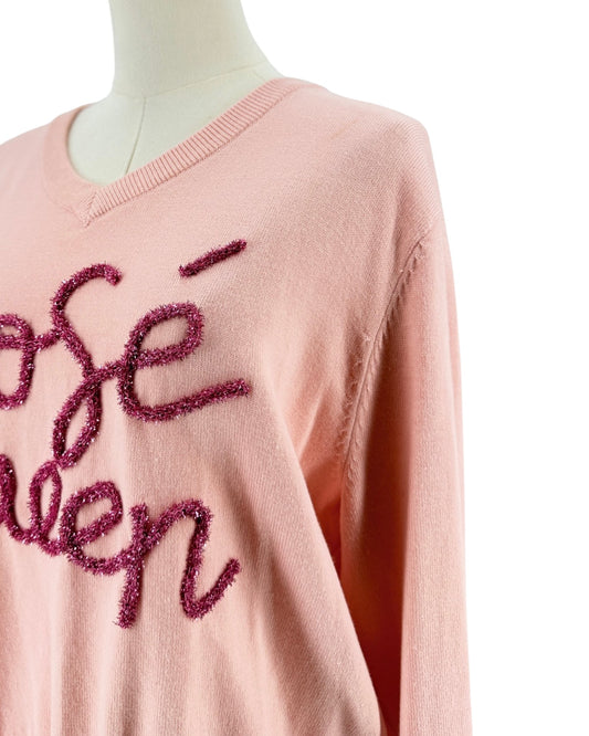 A close-up of a light pink knit sweater displayed on a mannequin, featuring textured, glittery magenta cursive lettering on the front.