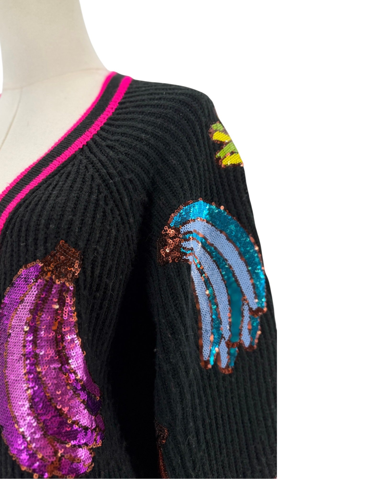 Close-up of a black ribbed knit sweater on a mannequin, featuring sequined banana motifs in purple, blue, and yellow, with a bright pink and black V-neck trim.