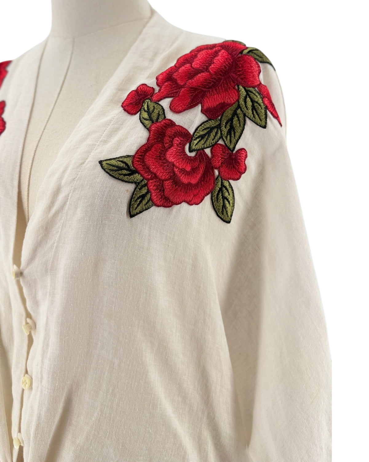 White 3/4 sleeve top with red and green embroidered flowers with a button front and bottom tie. on a mannequin on a white background