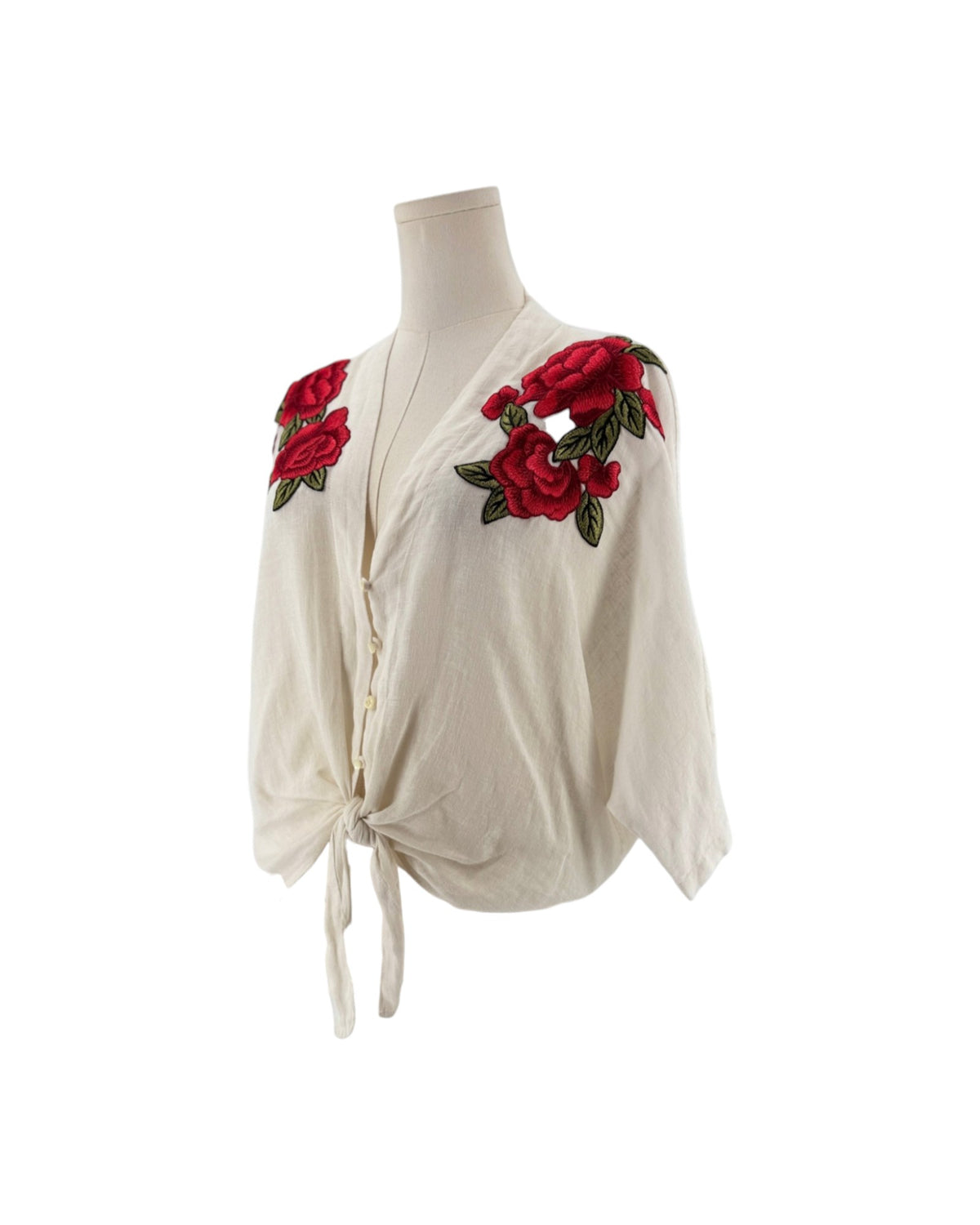 White 3/4 sleeve top with red and green embroidered flowers with a button front and bottom tie. on a mannequin on a white background