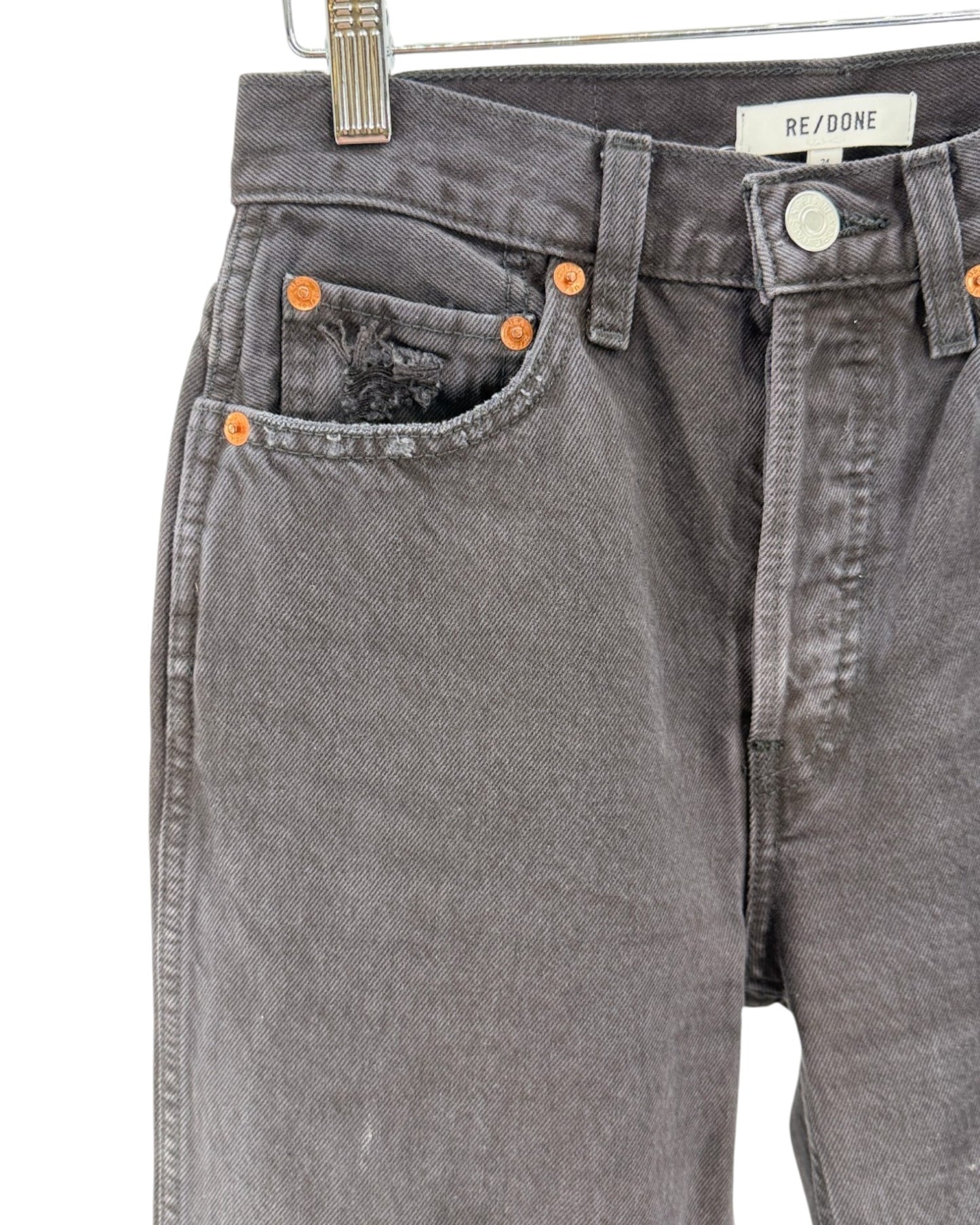 Close-up of a pair of faded charcoal gray denim jeans hanging on a metal hanger against a white background. The jeans feature orange copper-toned rivets, a distressed detail on the right pocket, and a white brand label reading 'REDONE' on the waistband.