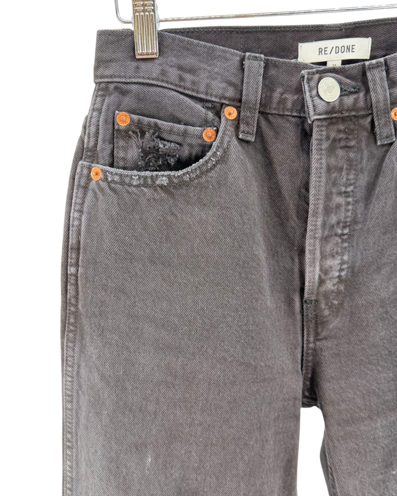 Close-up of a pair of faded charcoal gray denim jeans hanging on a metal hanger against a white background. The jeans feature orange copper-toned rivets, a distressed detail on the right pocket, and a white brand label reading 'REDONE' on the waistband.