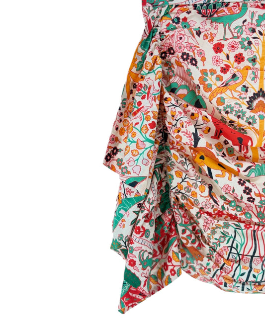 A close-up of a ruffled, asymmetrical skirt featuring a vibrant, multicolored animal and floral print on a cream background, set against a plain white studio backdrop.