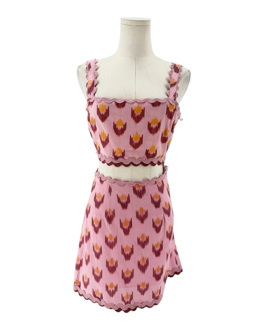 A pink sleeveless dress with a matching waist belt, displayed on a mannequin torso against a white background. The dress features a repeating ikat-style floral pattern in dark red and yellow, with scalloped edges.
