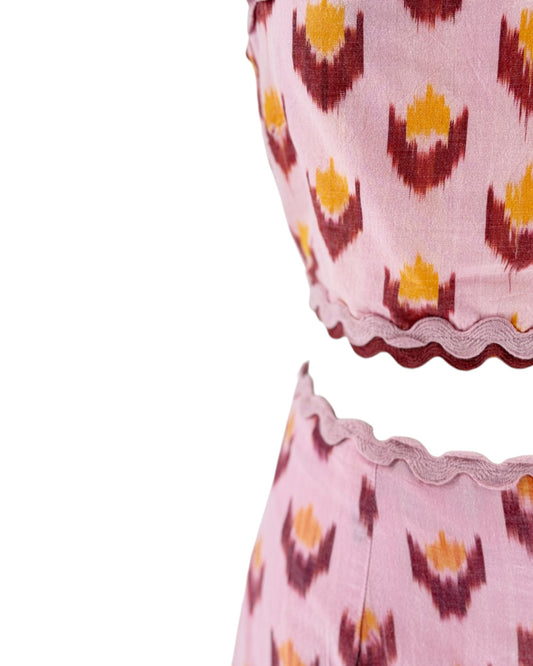 A close-up of a pink two-piece set featuring a matching crop top and shorts with a yellow and maroon ikat pattern, finished with scalloped lace trim.