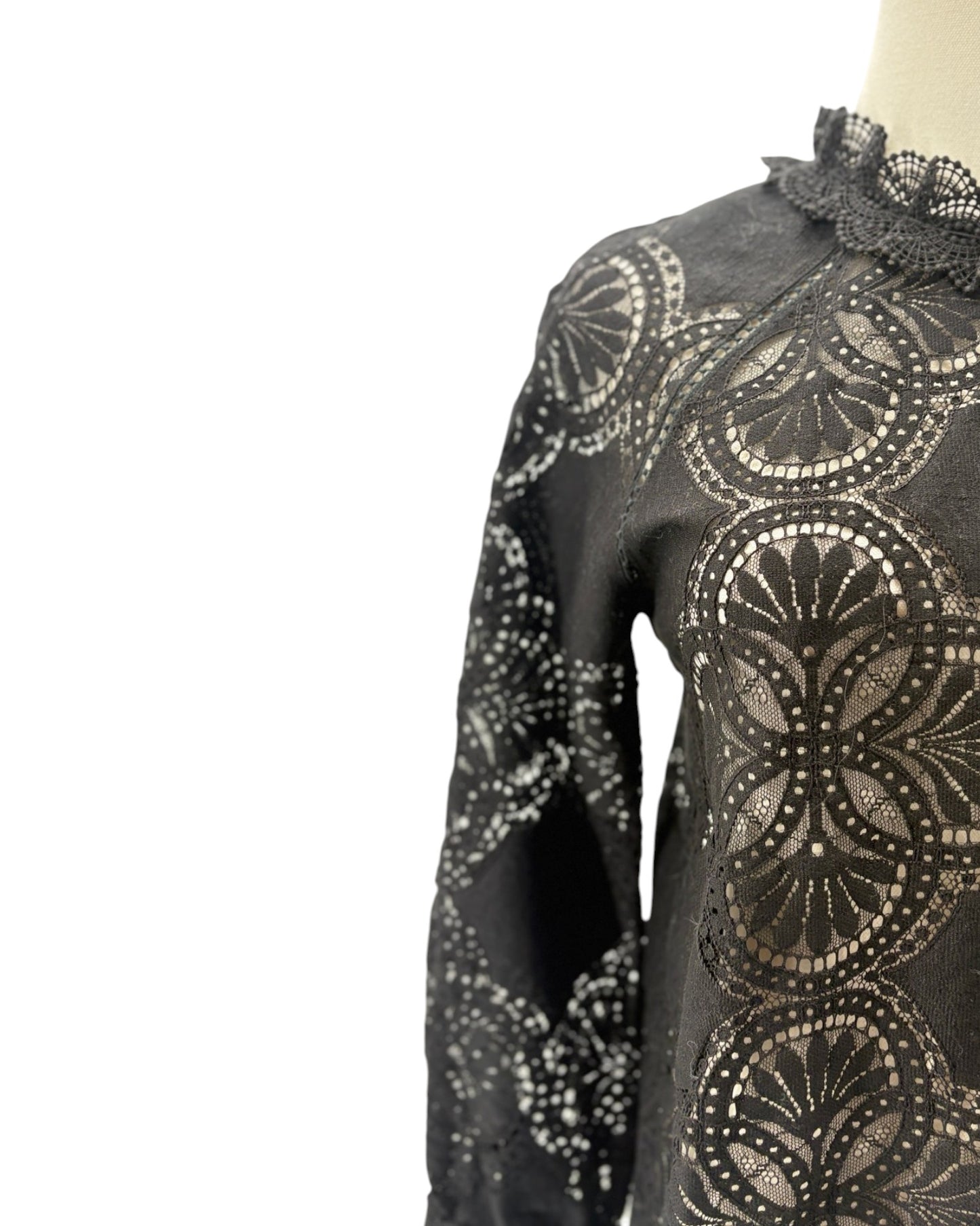 A close-up of a black long-sleeve blouse or dress featuring an intricate circular lace overlay pattern, displayed on a mannequin against a white background.
