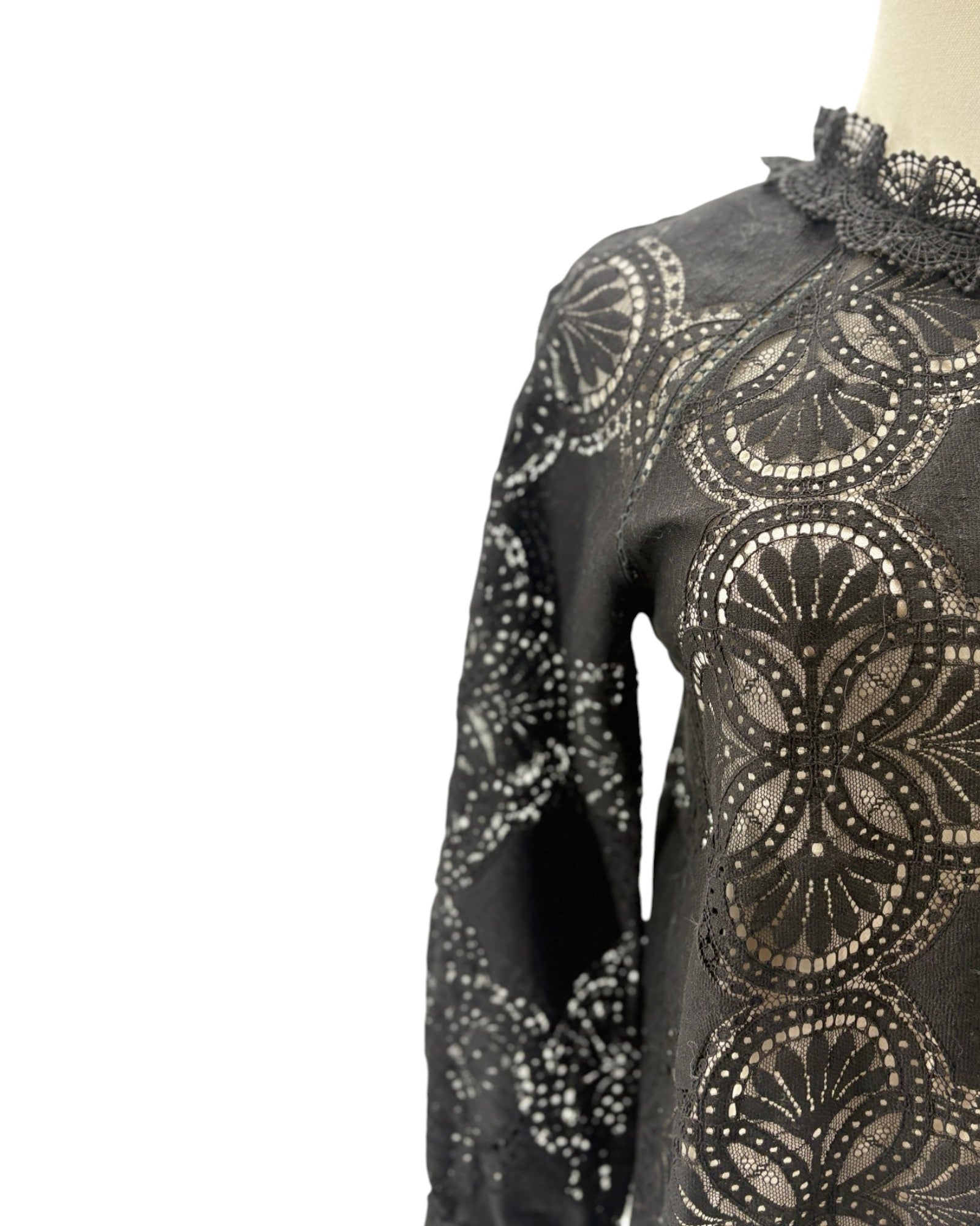 A close-up of a black long-sleeve blouse or dress featuring an intricate circular lace overlay pattern, displayed on a mannequin against a white background.