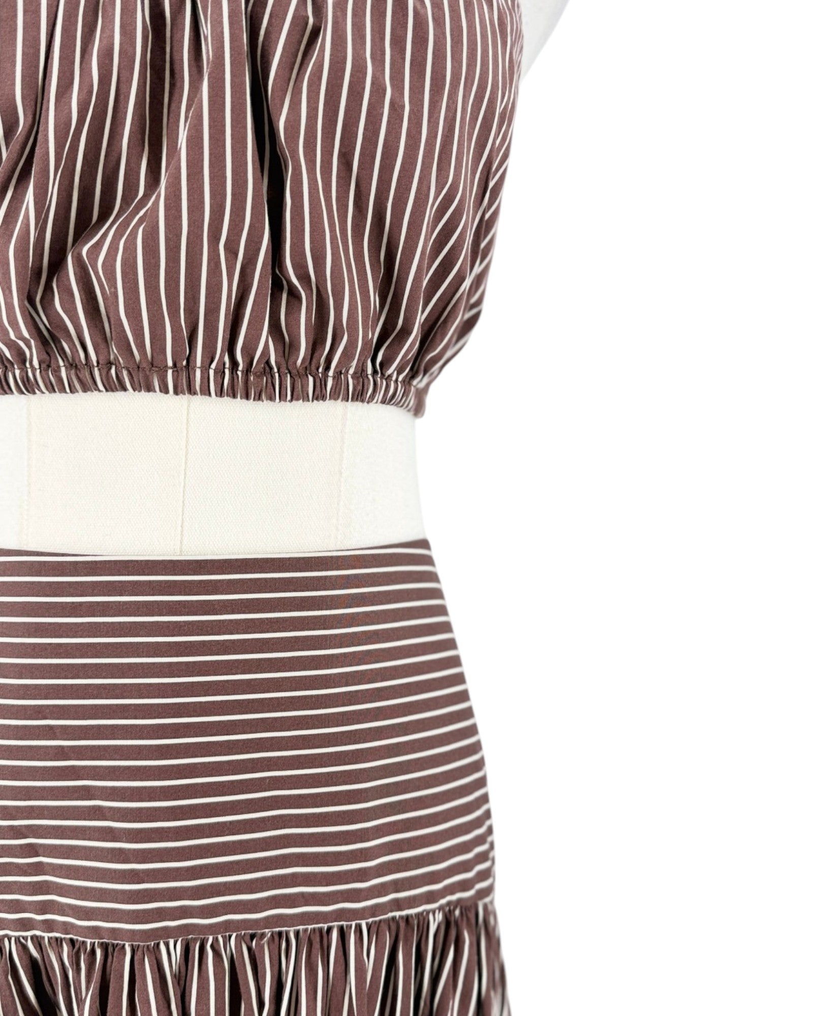 Close-up of a brown and white striped garment on a mannequin, showing a gathered waistband, a wide cream-colored waist panel, and a ruffled hem against a white background.