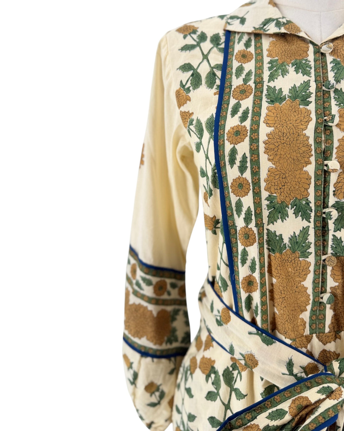 A close-up of a cream-colored dress with a green and brown floral pattern, featuring blue piping details and a buttoned front, displayed on a mannequin against a white background.