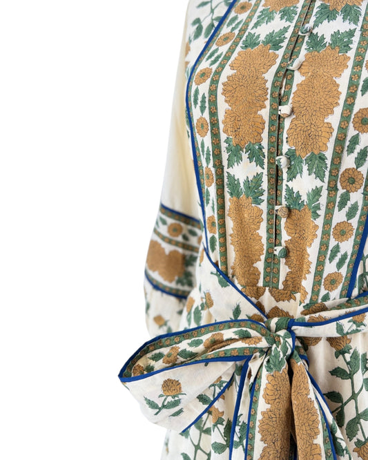 A close-up of a printed fabric garment or scarf with a cream base, large tan floral motifs, green leafy details, and blue edging, tied in a bow at the waist.