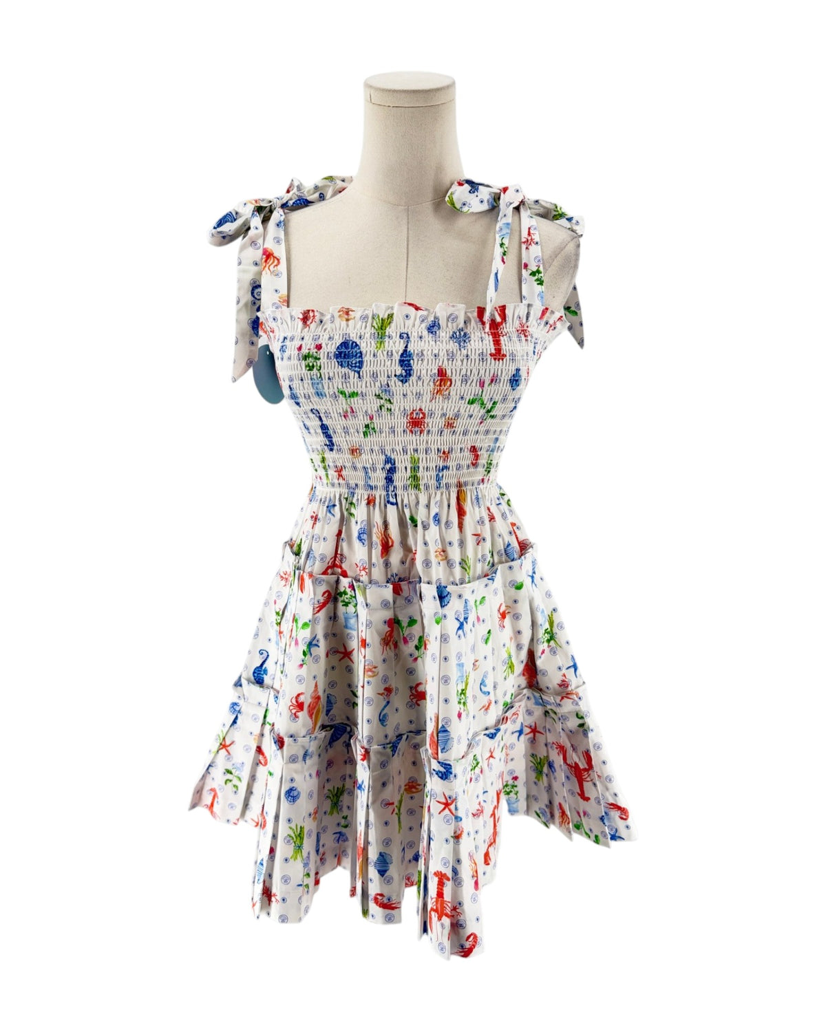 A sleeveless smocked dress with tie shoulder straps, featuring a colorful print of sea creatures like starfish, crabs, and fish on a white background, displayed on a mannequin torso against a white background.