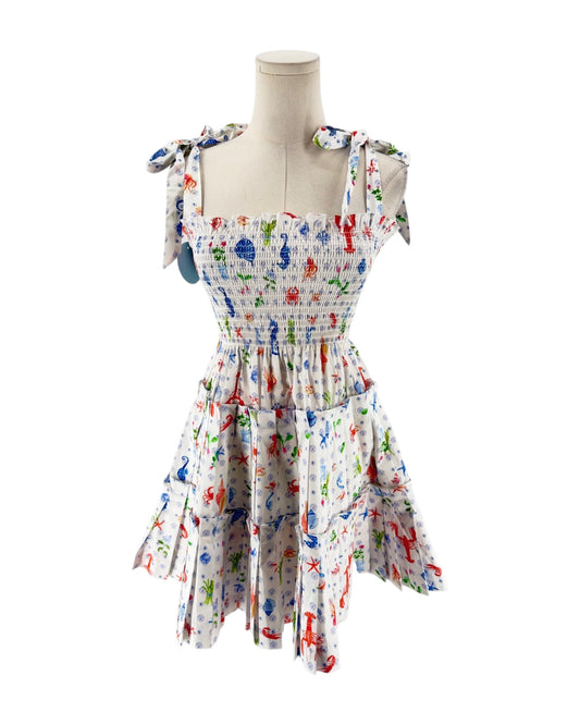 A sleeveless smocked dress with tie shoulder straps, featuring a colorful print of sea creatures like starfish, crabs, and fish on a white background, displayed on a mannequin torso against a white background.