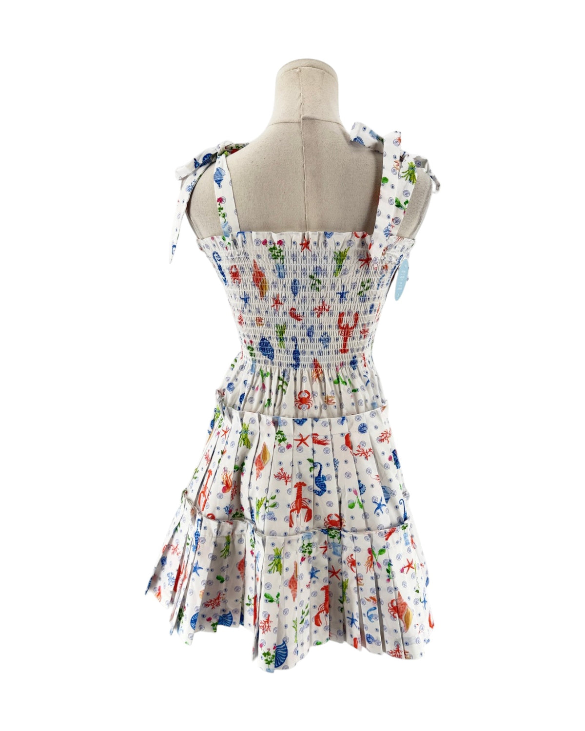 A sleeveless smocked dress with tie shoulder straps, featuring a colorful print of sea creatures like starfish, crabs, and fish on a white background, displayed on a mannequin torso against a white background.