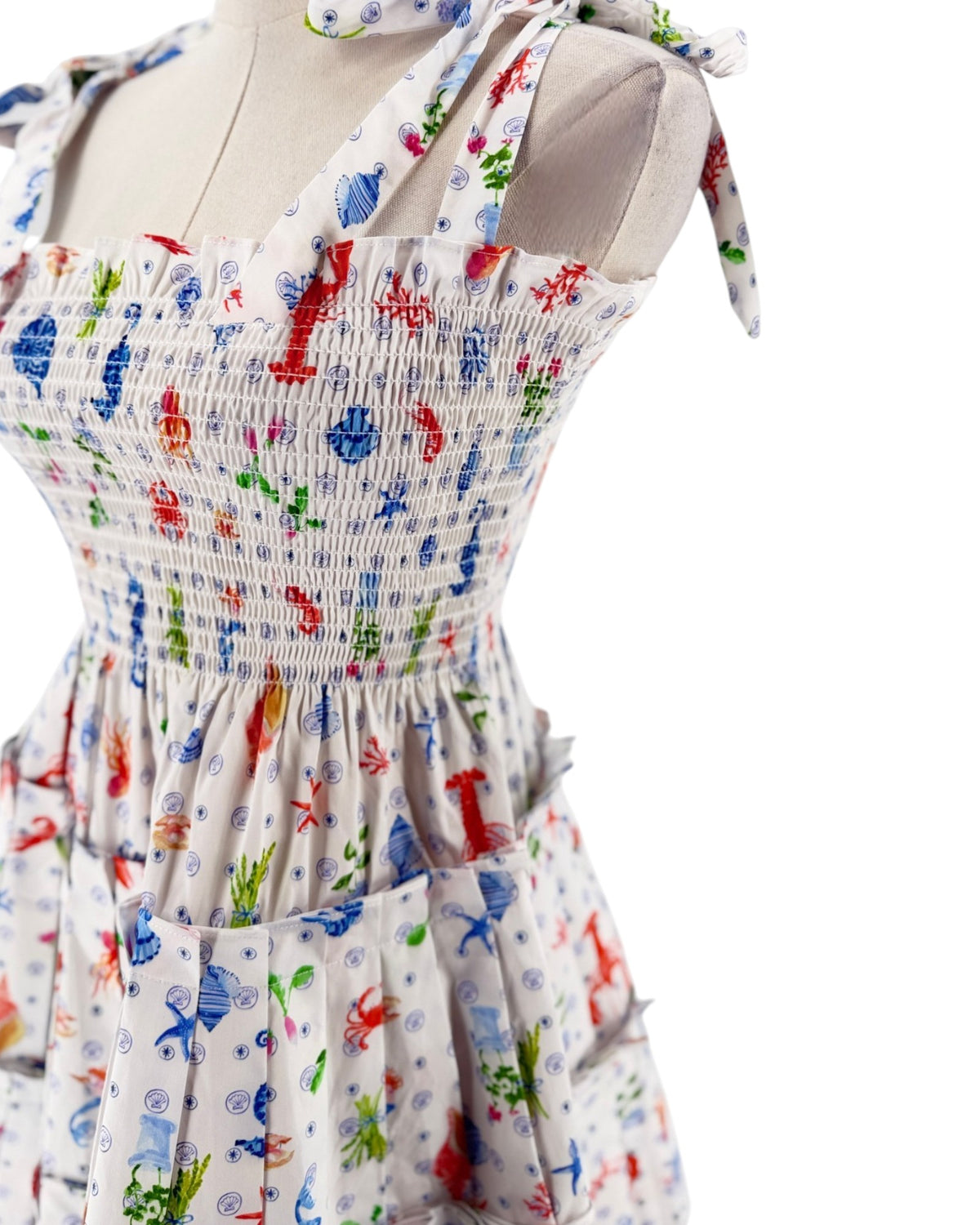 A sleeveless smocked dress with tie shoulder straps, featuring a colorful print of sea creatures like starfish, crabs, and fish on a white background, displayed on a mannequin torso against a white background.