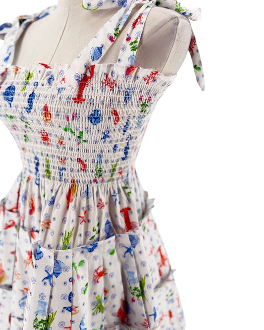A sleeveless smocked dress with tie shoulder straps, featuring a colorful print of sea creatures like starfish, crabs, and fish on a white background, displayed on a mannequin torso against a white background.