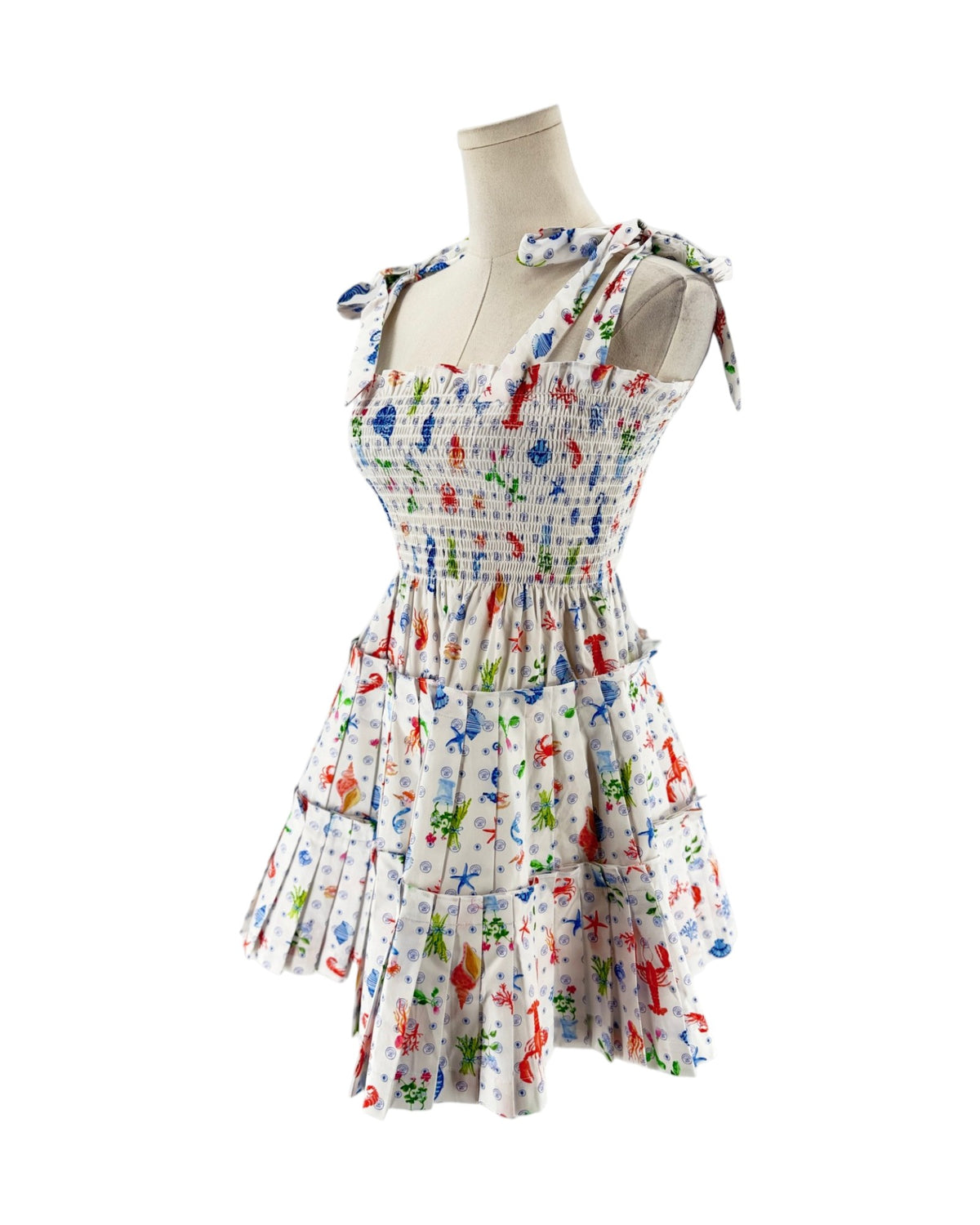 A sleeveless smocked dress with tie shoulder straps, featuring a colorful print of sea creatures like starfish, crabs, and fish on a white background, displayed on a mannequin torso against a white background.