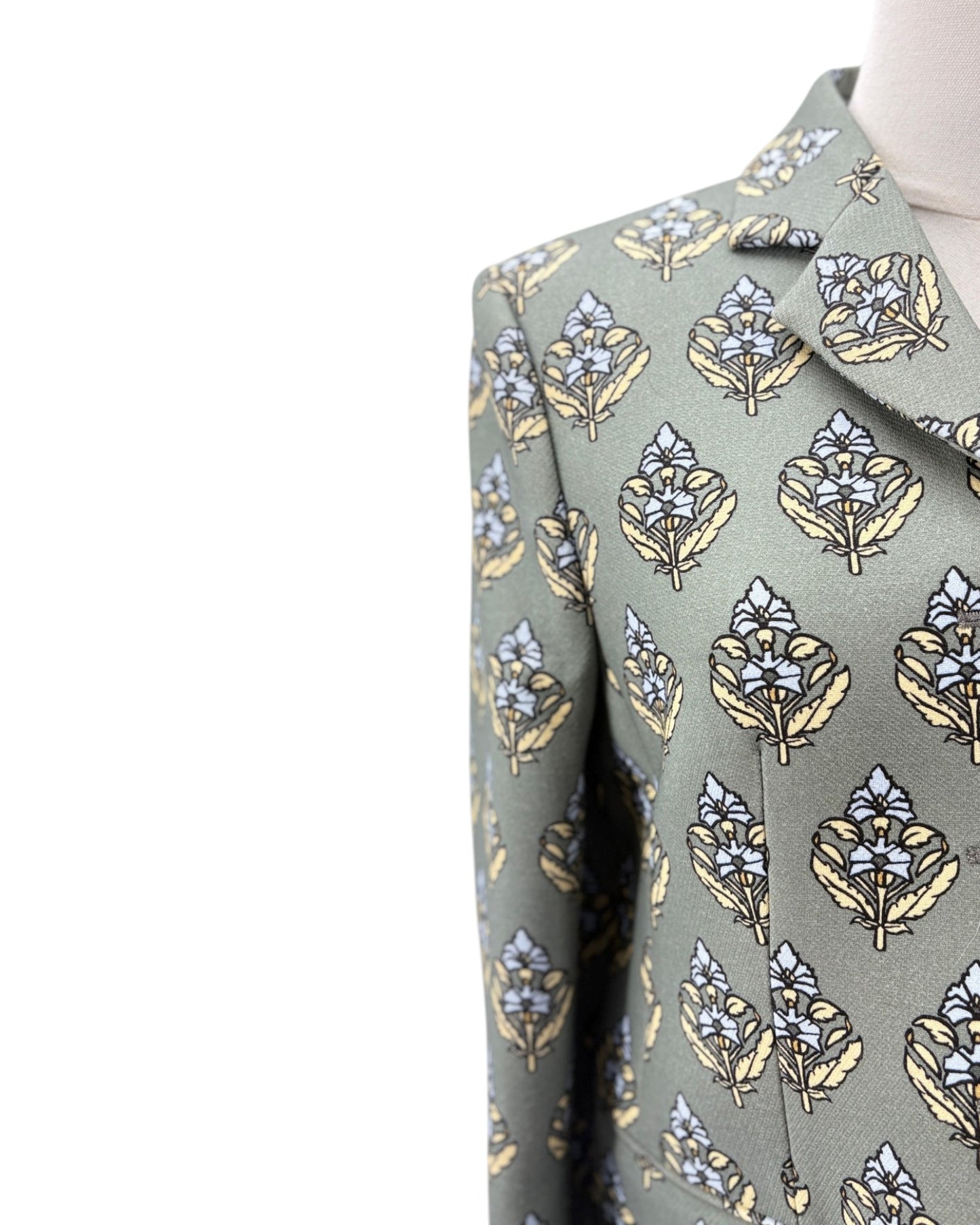 Close-up of a patterned suit jacket on a mannequin, featuring a sage green base with a repeating leaf and floral motif in beige, light blue, and white.