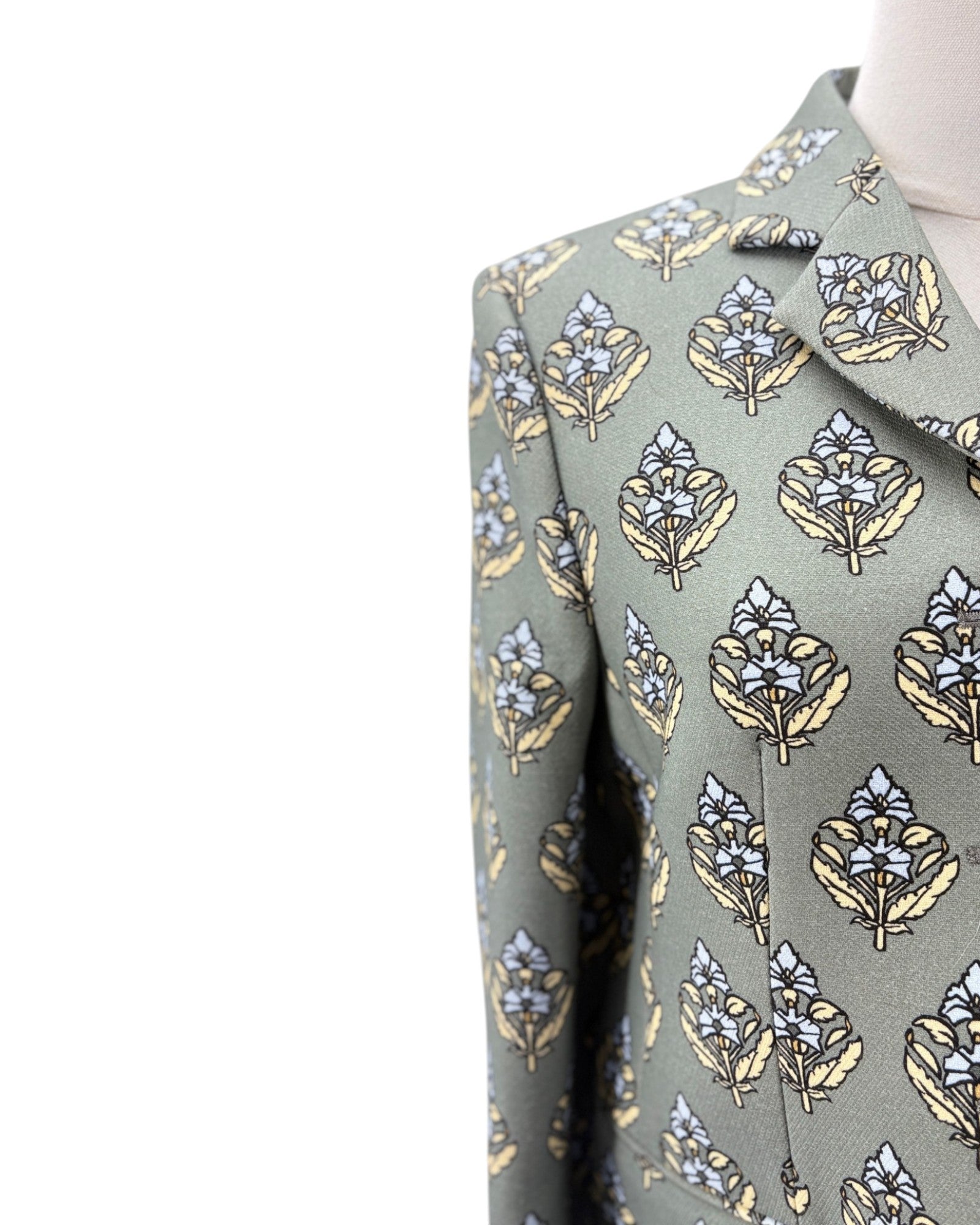 Close-up of a patterned suit jacket on a mannequin, featuring a sage green base with a repeating leaf and floral motif in beige, light blue, and white.