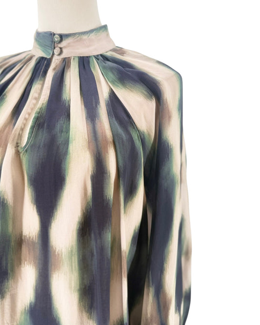 A close-up of a high-neck blouse displayed on a mannequin, featuring a pleated gathered bodice and a long sleeve. The fabric has an abstract, painterly pattern in beige, navy blue, and sage green.