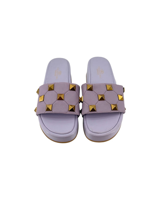 A pair of lavender slide sandals featuring a wide strap embellished with gold pyramid studs, shown from a top-down view on a white background.