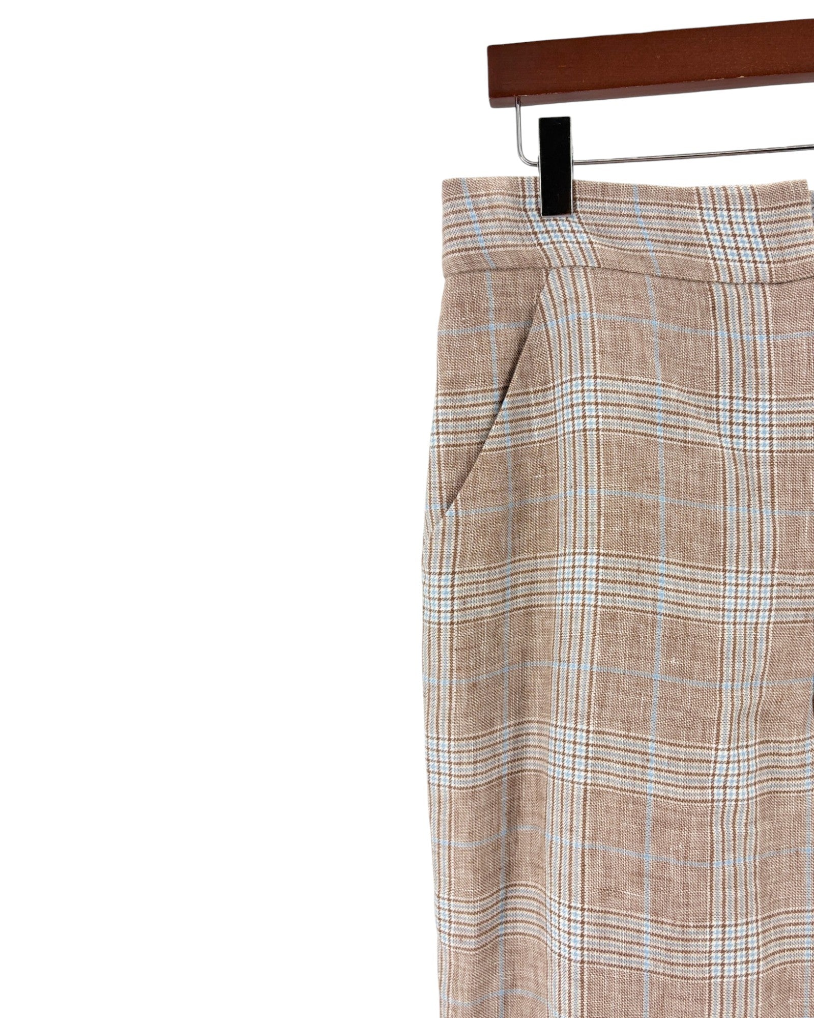 A close-up of a pair of plaid trousers hanging on a black hanger from a wooden hanger bar, set against a plain white background.